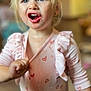 child, blonde_hair, blue_eyes, pink_shirt, heart_pattern, expression, indoor, person, portrait, toddler, clothing, hand, face, emotion, cute, young, baby, sleeve, rippled_fabric, background_blur