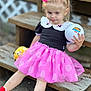 child, girl, pink_bow, pink_skirt, polka_dot, toy_ball, plush_toy, wooden_steps, outdoor, casual_clothing, red_socks, sitting, playful, cute, blonde_hair, portrait, daylight, young_child, expression, holding
