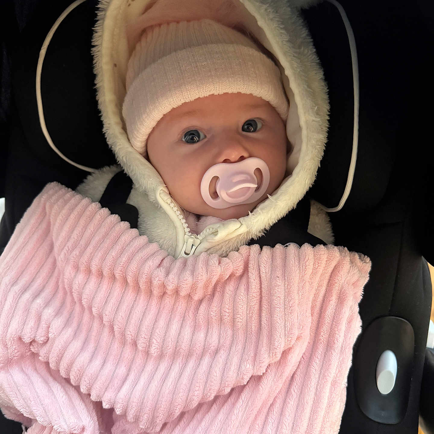 Polly-Mae is registered to the contest to win money with this photo: baby, blanket, car_seat, child, clothing, cozy, cute, face, hat, hood, indoors, infant, pacifier, person, pink, seated, soft_texture, warm, wide_eyes, wrapped