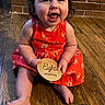 baby, child, infant, smiling, sitting, orange_dress, wooden_floor, brick_wall, holding_sign, eight_months, happy, indoors, person, cute, portrait, young_child, floor, home, playful, toddler