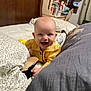 baby, smiling, bed, yellow_shirt, shoe, indoor, happy, person, furniture, books, pillow, blanket, wood, head, face, child, room, curtains, table, sitting