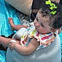baby, child, curly_hair, green_bow, striped_dress, baby_seat, blue_sheet, indoor, cute, portrait, skin, hands, feet, earring, infant, sitting, colorful_clothing, natural_light, curious, soft_texture