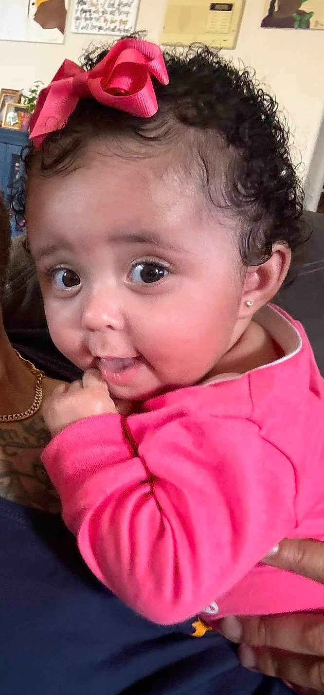 Nyrobi joined the competition — help win amazing prizes! baby, child, pink_clothing, curly_hair, bow, earrings, indoor, person, cute, smiling, hand, face, eyes, infant, portrait, holding, home, couch, skin, young