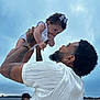 Nyrobi joined the competition — help win amazing prizes! baby, man, tattoo, watch, curly_hair, bow, white_clothing, outdoor, sky, water, portrait, holding, child, beard, earring, glasses, facial_hair, person, family, love