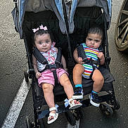 Nyrobi is registered to the contest to win money with this photo: toddler, double_stroller, child, parking_lot, asphalt, baby, footwear, harness, pink_bows, floral_outfit, striped_onesie, expression, curious, serious, black_harness, sunlight, outdoor, wheels, side_by_side, cute