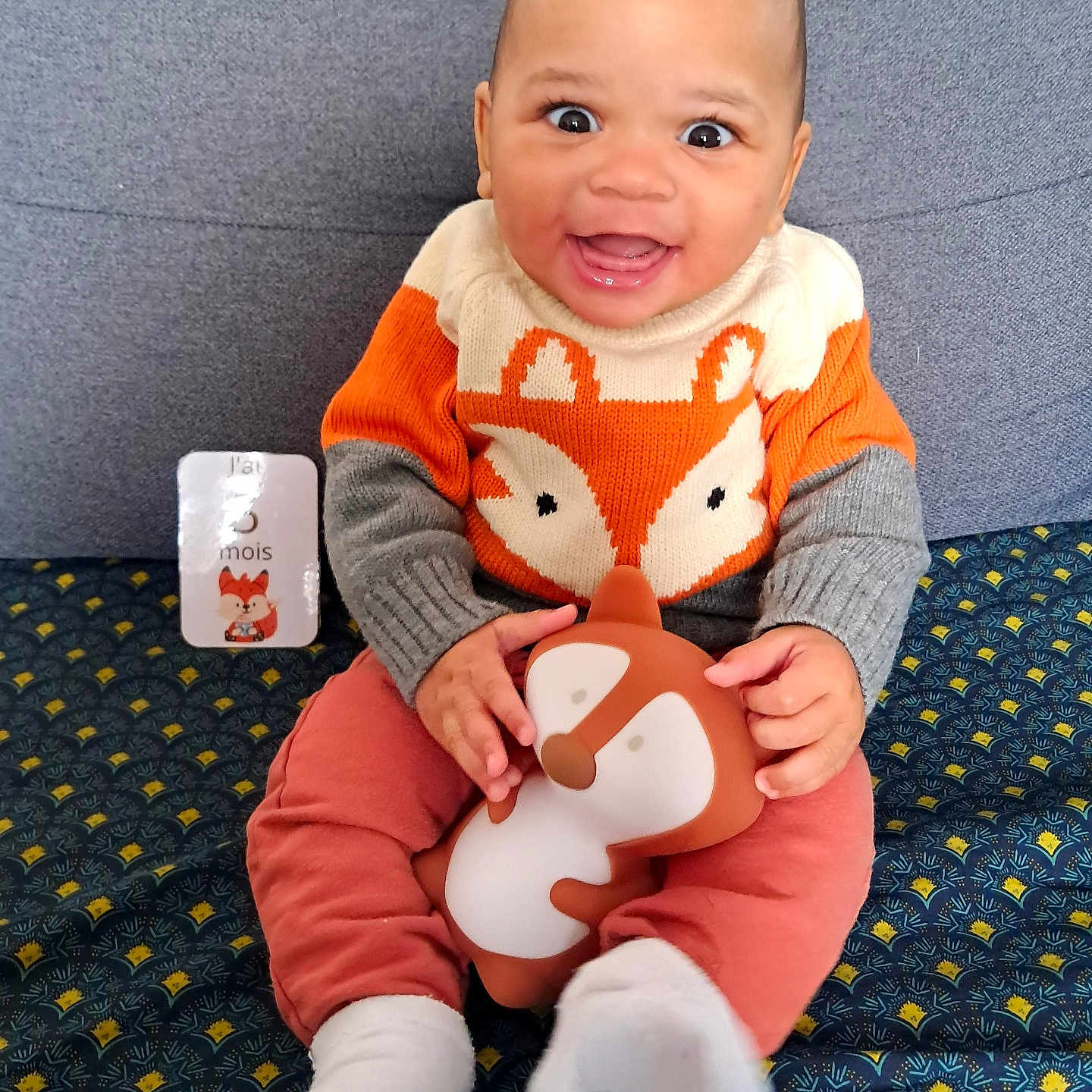 Lyssandro participe au concours pour gagner de l'argent avec cette photo : baby, child, smiling, fox_sweater, toy, stuffed_animal, sitting, cushion, patterned_fabric, white_socks, happy, indoor, portrait, cute, clothing, person, infant, playful, bright_colors, closeup