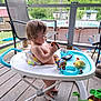 Jordan is registered to the contest to win money with this photo: toddler, baby_walker, toy_car, rubiks_cube, plush_toy, outdoor, porch, wooden_floor, child, summer, pool, greenery, chair, playtime, cute, curious, daylight, casual, footwear, relaxing