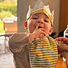 Lucas a rejoint le concours — aidez-le/la à gagner de superbes lots ! baby, bib, bokeh, child, crown, cute, dining_table, eyelashes, eyes, finger_in_mouth, hand, indoor, infant, paper_crown, parent_hand, portrait, smile, striped_bib, warm_lighting, wooden_table
