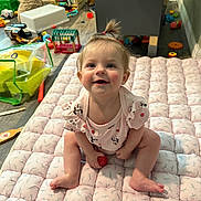Charlie is registered to the contest to win money with this photo: baby, child, indoor, playroom, toy, smiling, person, mat, floor, hair, pony_tail, cute, clothing, pink, sitting, face, expression, furniture, books, colorful