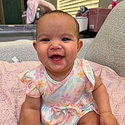 Oaklynn is registered to the contest to win money with this photo: baby, child, smiling, floral_dress, pink_blanket, sitting, indoors, face, happy, cute, infant, person, home, soft_light, window_blinds, plush, arm, head, skin, furniture