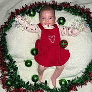 Rosie joined the competition — help win amazing prizes! baby, infant, red_dress, christmas_ornaments, green_baubles, tinsel, holiday_decor, white_blanket, festive, smiling, happy, cute, lying_down, holiday_theme, face, person, christmas, seasonal, indoors, celebration