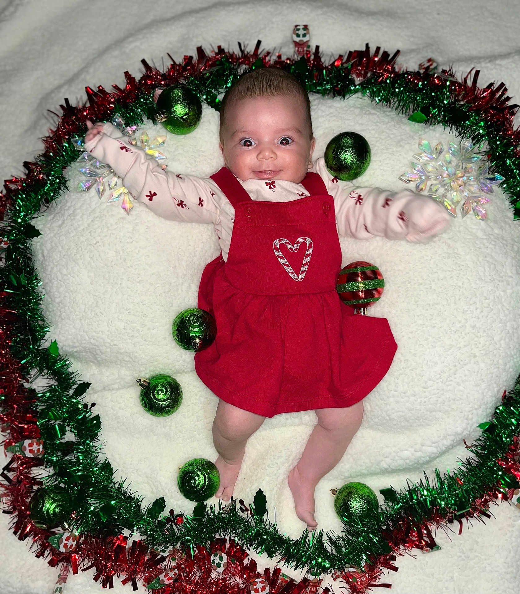 Rosie joined the competition — help win amazing prizes! baby, infant, red_dress, christmas_ornaments, green_baubles, tinsel, holiday_decor, white_blanket, festive, smiling, happy, cute, lying_down, holiday_theme, face, person, christmas, seasonal, indoors, celebration