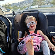 Kaylee is registered to the contest to win money with this photo: toddler, child, car_seat, glasses, blanket, foot, smile, happy, car_interior, seatbelt, clothing, person, window, mountains, outdoor_view, toy, pink, silver_pants, vehicle, rear_window