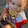 blue_eyes, casual_clothing, child, cracker, curly_hair, face, food, hand, happy, marshmallow, messy, outdoor, portrait, red_shirt, shorts, smore, snack, summer, sunlight, toddler