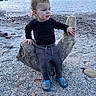beach, black_sweater, blue_shoes, casual_clothing, child, curly_hair, daytime, driftwood, gray_pants, nature, outdoor, playful, portrait, rocks, sand, scenic, sea, small_child, toddler, water