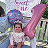 baby, child, birthday, first_birthday, balloon, number_1, party, celebration, outdoor, smiling, happy, chair, cushion, banner, decoration, sunlight, ribbon, clothing, footwear, person