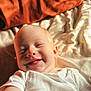baby, bed, blanket, child, cozy, cute, face, happy, head, indoor, infant, lying_down, orange_blanket, portrait, skin, smiling, soft_light, tongue, warm_colors, white_clothing