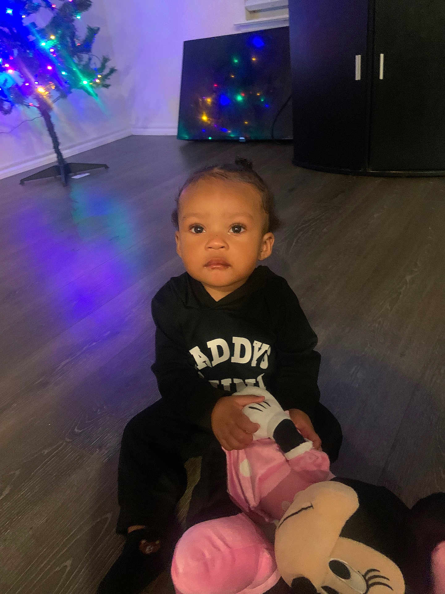 Taylen is registered to the contest to win money with this photo: toddler, child, plush_toy, minnie_mouse, black_hoodie, indoor, wooden_floor, christmas_tree, colorful_lights, toy, sitting, young_child, face, person, holiday, reflection, furniture, home, cute, expression
