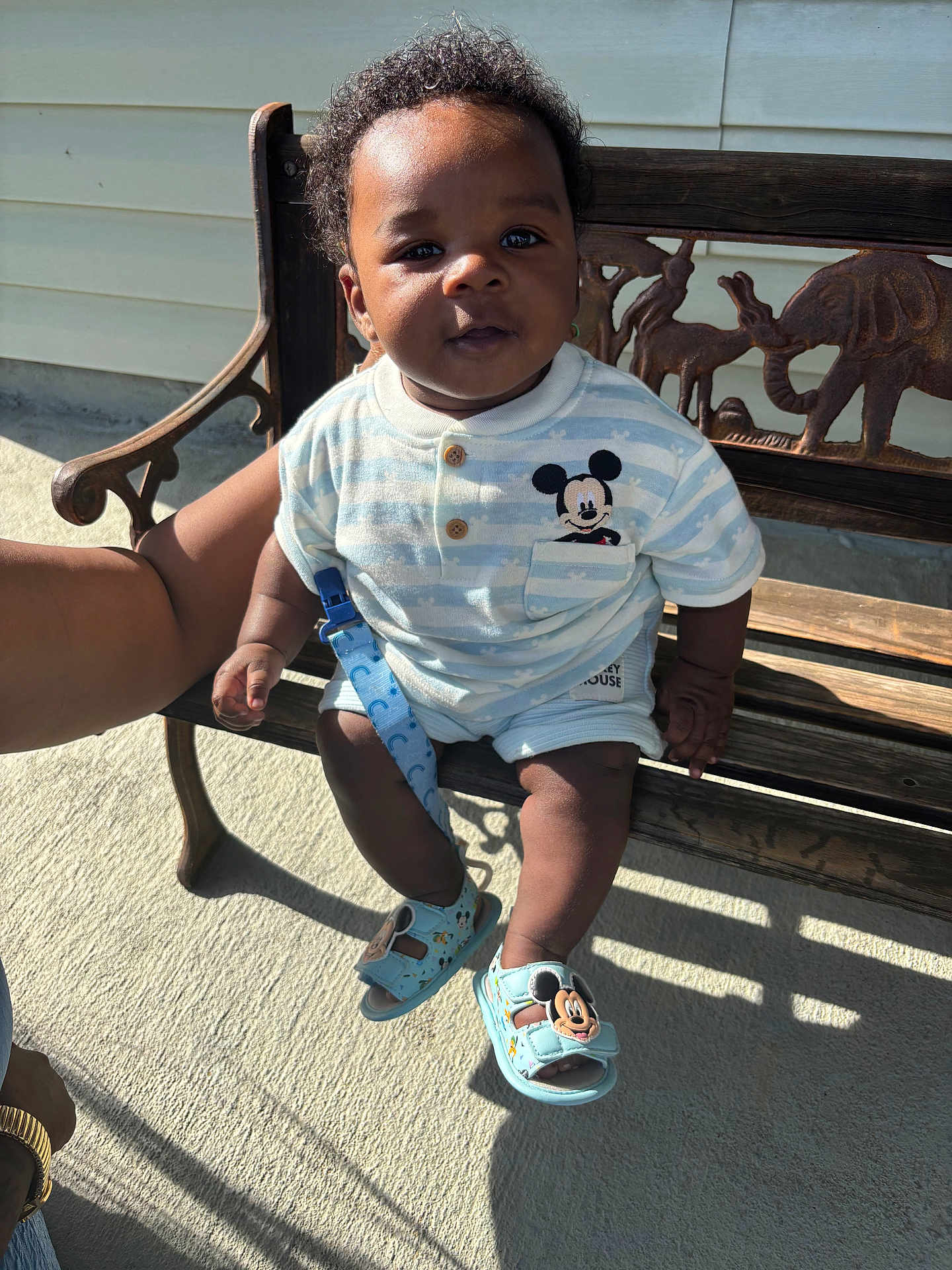 Kycen is registered to the contest to win money with this photo: baby, child, bench, wood, carving, mickey_mouse, clothing, sandals, outdoor, sunlight, shadow, person, cute, curious, smile, arm, hand, footwear, face, happy