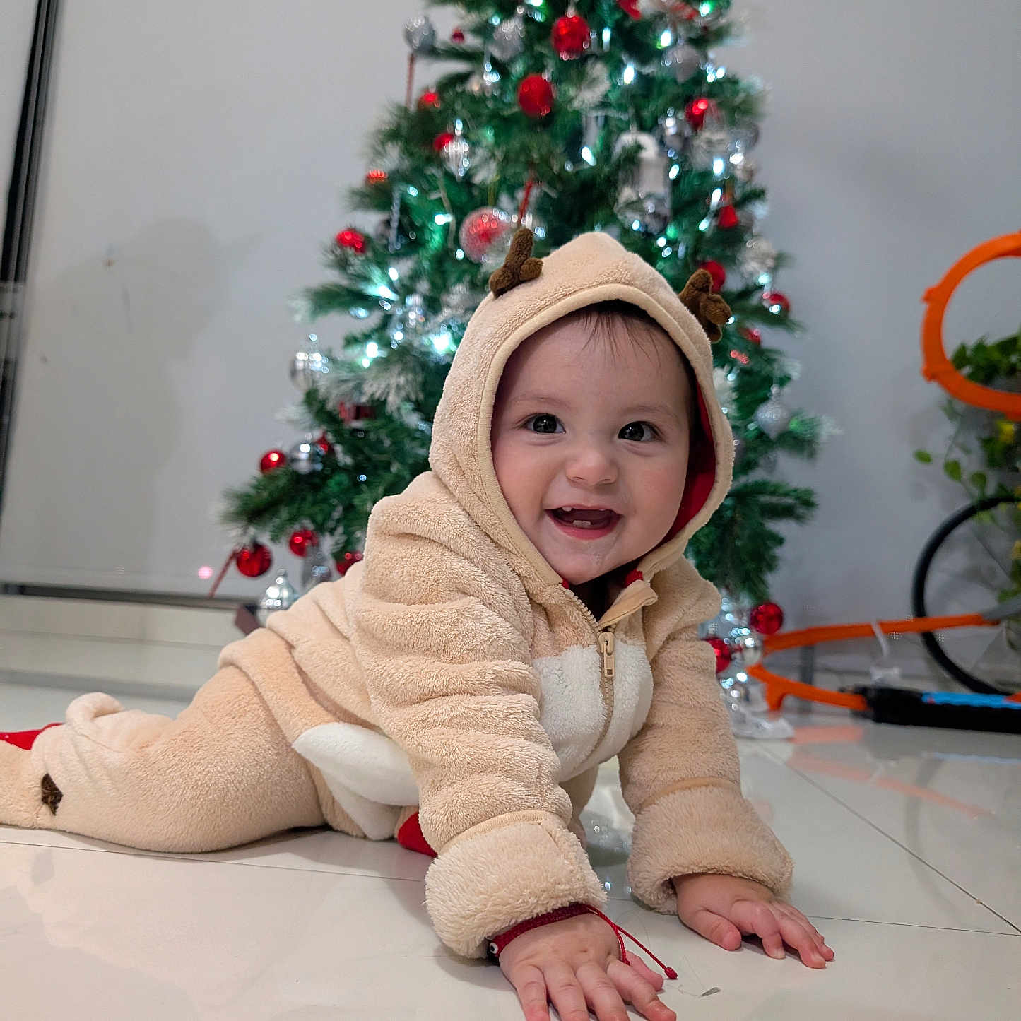 David is registered to the contest to win money with this photo: baby, child, crawling, reindeer_onesie, holiday, christmas_tree, decorations, indoor, smiling, happy, festive, floor, toy, cute, infant, holiday_lights, seasonal, warm_clothing, joyful, portrait