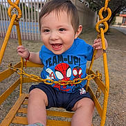 David is registered to the contest to win money with this photo: blue_shirt, child, daylight, fence, grass, hand, happy, metal, outdoor, park, person, playground, shorts, smiling, socks, spiderman, swing, toddler, tree, yellow_swing