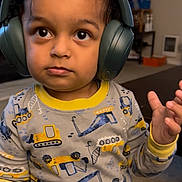 Addie joined the competition — help win amazing prizes! child, toddler, headphones, pajamas, construction_vehicles, indoor, carpet, curly_hair, big_eyes, long_sleeves, yellow, gray, playful, cute, expression, person, young_child, living_room, furniture, hand