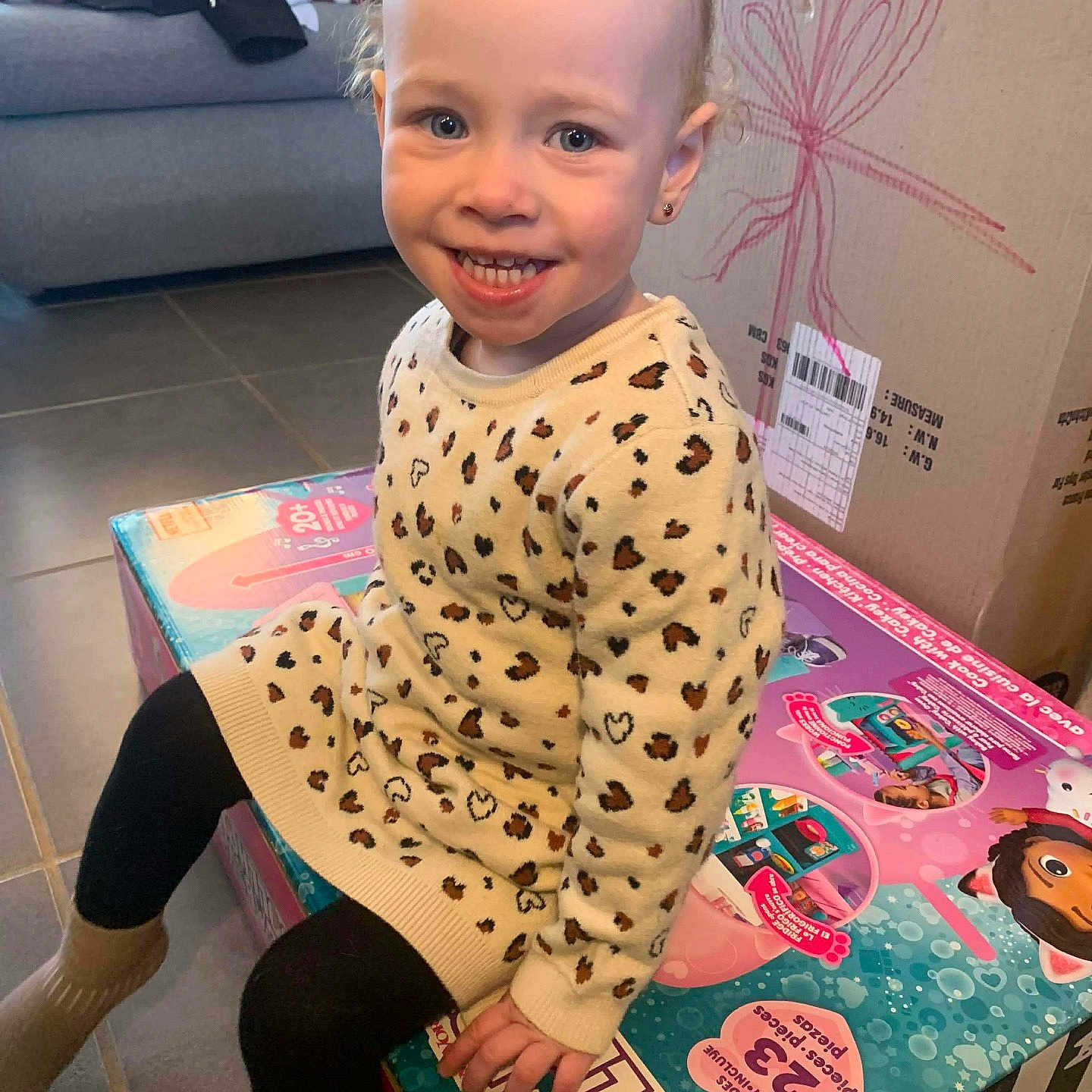 Andréa a rejoint le concours — aidez-le/la à gagner de superbes lots ! blond_hair, blue_eyes, cardboard_box, child, couch, cute, earring, floor_tiles, happy, indoor, leggings, leopard_print_sweater, living_room, portrait, present, sitting, smiling, socks, toddler, toy_box