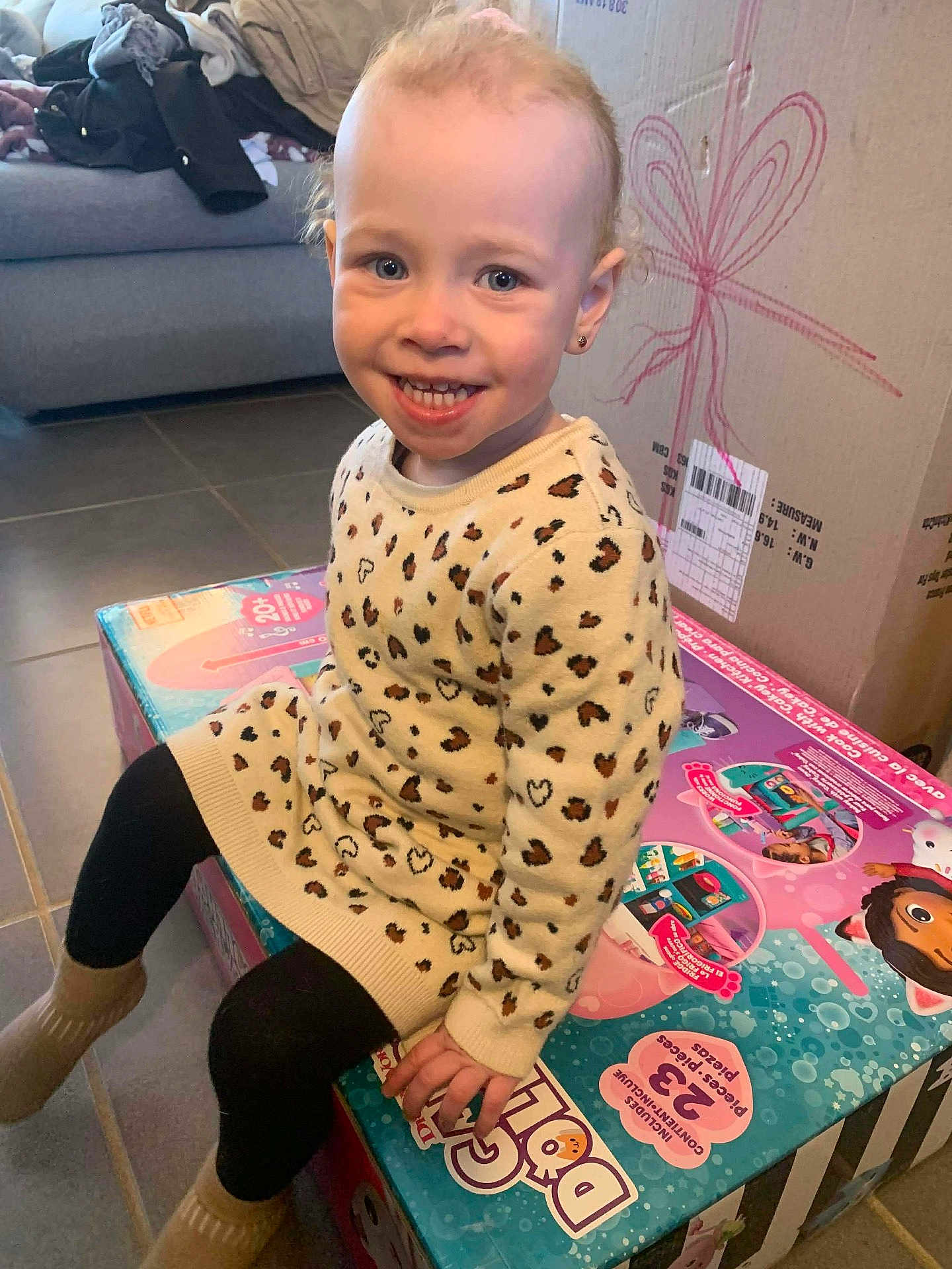 Andréa a rejoint le concours — aidez-le/la à gagner de superbes lots ! child, toddler, smiling, leopard_print_sweater, sitting, toy_box, cardboard_box, living_room, couch, floor_tiles, blond_hair, blue_eyes, earring, leggings, socks, present, portrait, happy, indoor, cute