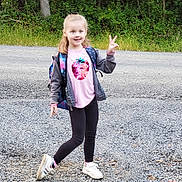 Ava is registered to the contest to win money with this photo: child, girl, outdoor, gravel, peace_sign, backpack, pink_shirt, sequins, strawberry, jacket, leggings, sneakers, smile, posing, nature, greenery, road, playful, happy, casual_clothing