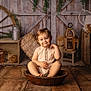 toddler, child, smile, wooden_bowl, wooden_floor, rustic, vintage, wagon_wheel, hay_bale, watering_can, lantern, wooden_crate, plant, indoor, portrait, cute, sitting, cozy, farm_style, decor