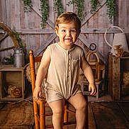 Loïs a rejoint le concours — aidez-le/la à gagner de superbes lots ! toddler, child, smiling, sitting, wooden_chair, romper, barefoot, indoor, rustic, farmhouse, wood_floor, greenery, decor, vintage, happy, portrait, person, cute, warm_lighting, cozy