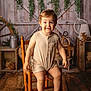toddler, child, smiling, sitting, wooden_chair, romper, barefoot, indoor, rustic, farmhouse, wood_floor, greenery, decor, vintage, happy, portrait, person, cute, warm_lighting, cozy