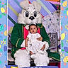Lylah joined the competition — help win amazing prizes! baby, child, person, mascot, easter_bunny, costume, glasses, green_coat, bench, indoor, portrait, holiday, easter_theme, decorations, ribbon, year_2026, floral_pattern, colorful_background, white_dress, festive