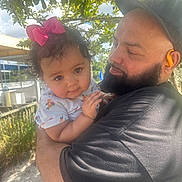 Aria is registered to the contest to win money with this photo: baby, child, man, holding, outdoor, tree, sunlight, beard, hat, pink_bow, curly_hair, nature, affection, portrait, casual_clothing, closeup, daylight, smiling, family, gentle