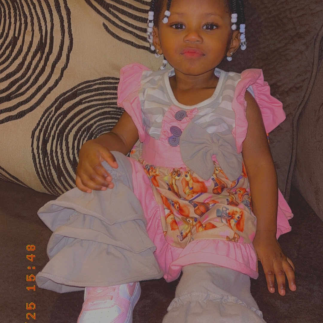 Amirica is registered to the contest to win money with this photo: beads, braided_hair, child, couch, cozy, cute, fashion, girl, home, indoor, patterned_pillow, pillow, pink_clothing, portrait, relaxed, ruffled_pants, seated, smiling, sneakers, toddler