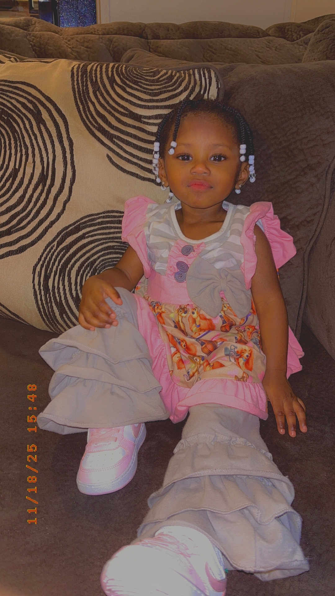 Amirica is registered to the contest to win money with this photo: toddler, child, girl, braided_hair, beads, pink_clothing, ruffled_pants, sneakers, couch, pillow, patterned_pillow, indoor, cozy, smiling, cute, fashion, seated, portrait, home, relaxed