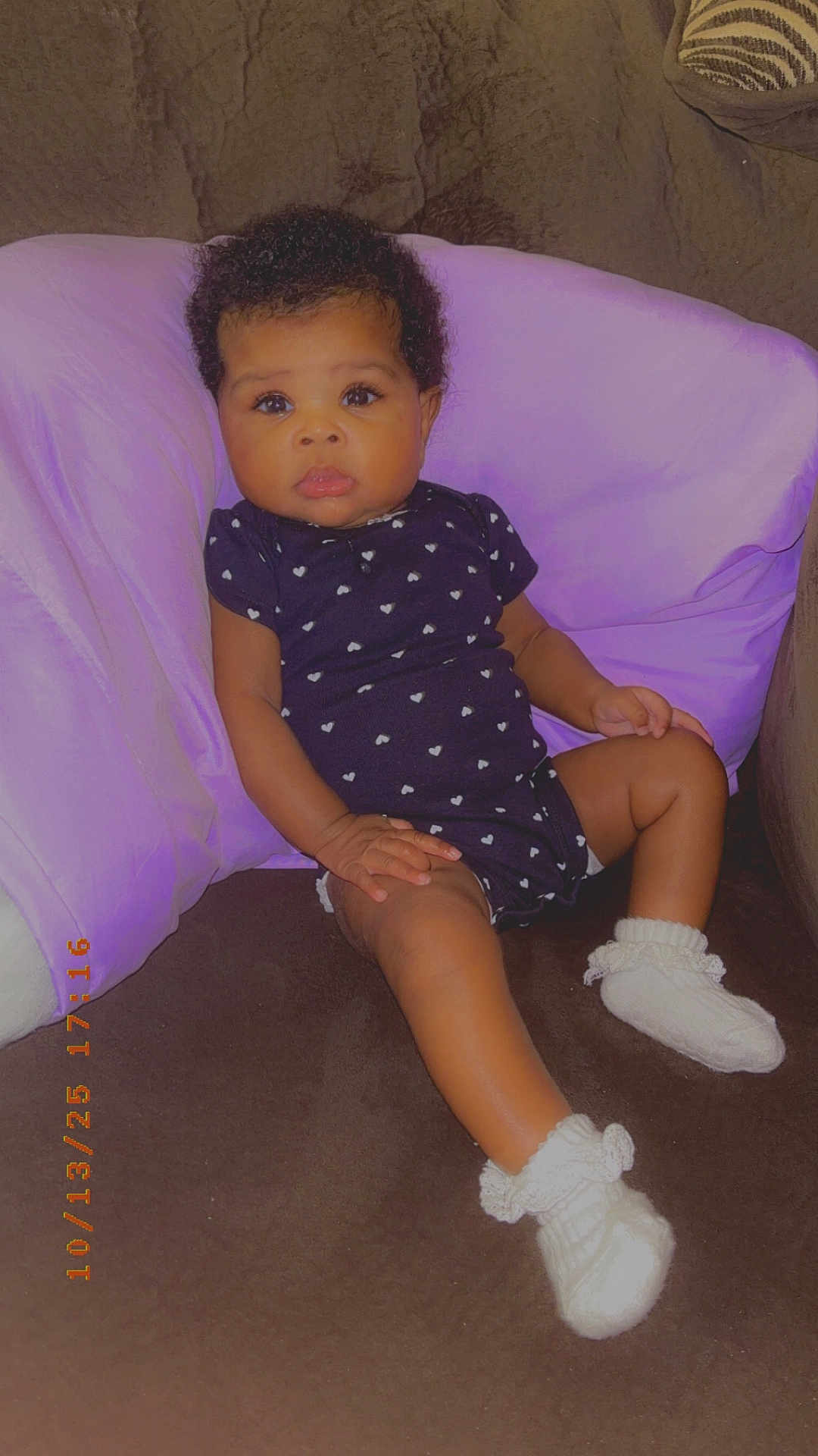 Ava is registered to the contest to win money with this photo: baby, brown_furniture, child, couch, cozy, curly_hair, cute, heart_pattern, indoor, infant, knitted_booties, looking_at_camera, onesie, portrait, purple_cushion, relaxed, sitting, small_hands, soft_lighting, white_socks