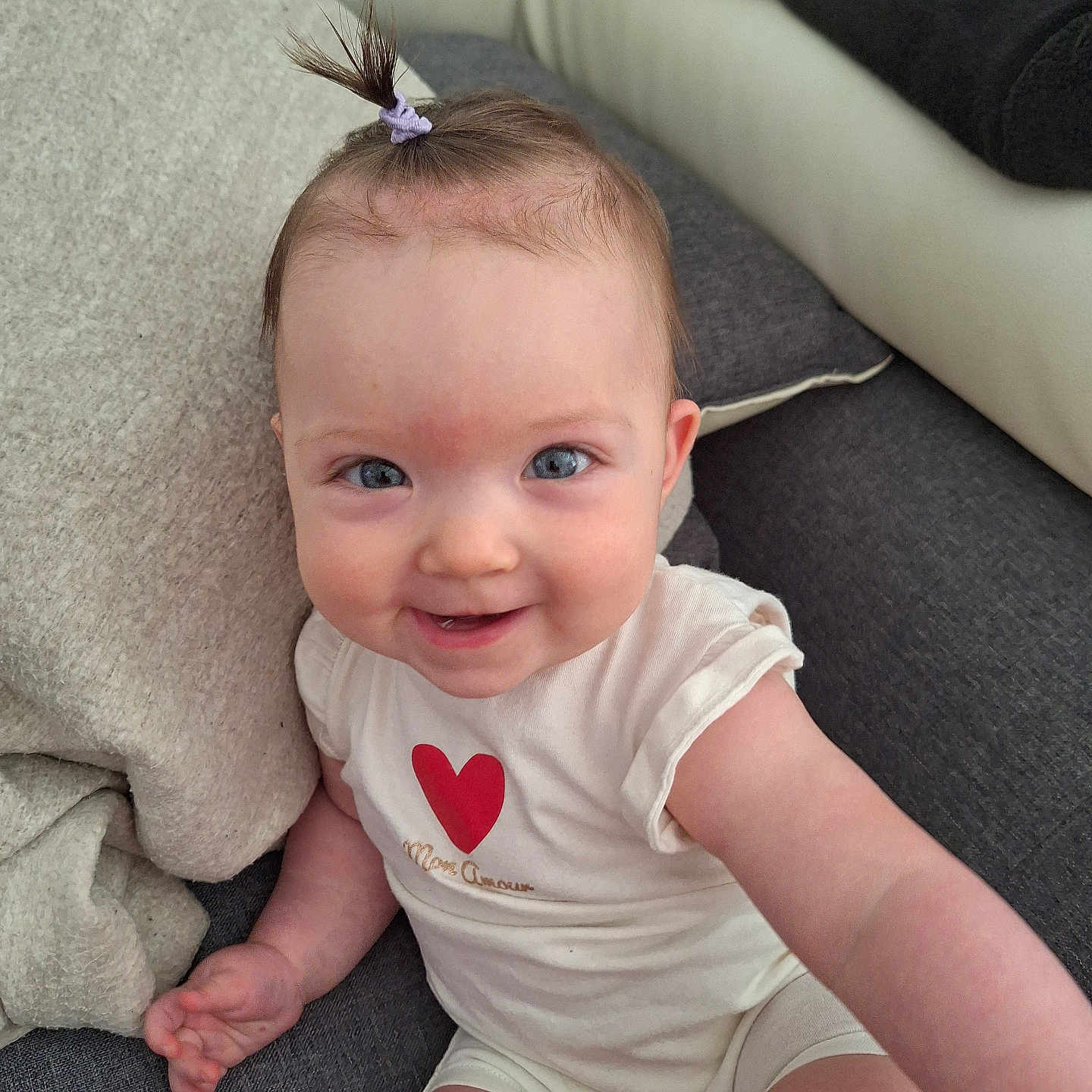 Eléna participe au concours pour gagner de l'argent avec cette photo : baby, blue_eyes, casual, child, couch, cute, face, hair_tie, hand, happy, indoor, infant, person, pillow, relaxed, sitting, skin, smiling, white_clothing, young_child