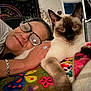 person, cat, siamese_cat, glasses, ring, bracelet, blanket, floral_pattern, indoor, close_up, smile, face, pet, animal, cozy, home_decor, tapestry, heart_decoration, hand, resting