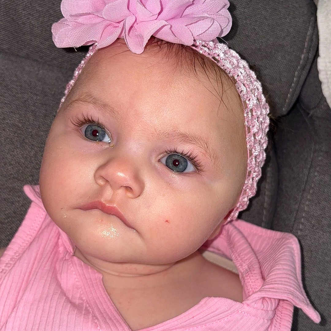 Jeanelle a rejoint le concours — aidez-le/la à gagner de superbes lots ! baby, blue_eyes, child, close_up, cushion, cute, expression, face, fashion, flower, gray_background, headwear, indoor, infant, long_lashes, pink_headband, pink_outfit, portrait, sitting, skin