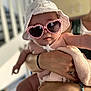 baby, child, sunglasses, hat, white_hat, heart_shaped_glasses, blanket, pink_blanket, arm, hand, person, indoor, blurred_background, cute, fashion, accessory, holding, skin, closeup, portrait
