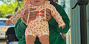 Camëlia a rejoint le concours — aidez-le/la à gagner de superbes lots ! baby, child, person, hood, clothing, striped_shorts, green_hoodie, outdoor, sunlight, trees, hand, face, expression, blurred_background, table, snack_bag, window, building, legs, holding