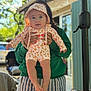 baby, child, person, hood, clothing, striped_shorts, green_hoodie, outdoor, sunlight, trees, hand, face, expression, blurred_background, table, snack_bag, window, building, legs, holding