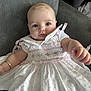 baby, child, dress, white_dress, pink_embroidery, couch, indoor, portrait, cute, infant, young_child, reaching_hand, blue_eyes, face, person, sitting, soft_lighting, furniture, close_up, adorable