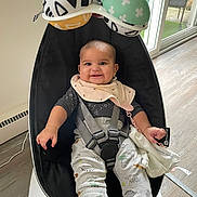 Serine is registered to the contest to win money with this photo: baby, smiling, baby_rocker, bib, toy, hanging_toy, indoor, floor, window, natural_light, clothing, dinosaur_pattern, child, seat_belt, black_seat, white_base, happy, person, infant, modern_furniture
