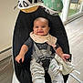 baby, smiling, baby_rocker, bib, toy, hanging_toy, indoor, floor, window, natural_light, clothing, dinosaur_pattern, child, seat_belt, black_seat, white_base, happy, person, infant, modern_furniture