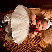 Delanis is registered to the contest to win money with this photo: baby, infant, child, dress, headband, blanket, pillow, brown_background, white_dress, red_top, black_shoes, white_socks, portrait, laying_down, cute, indoors, three_months, decor, soft_texture, cozy