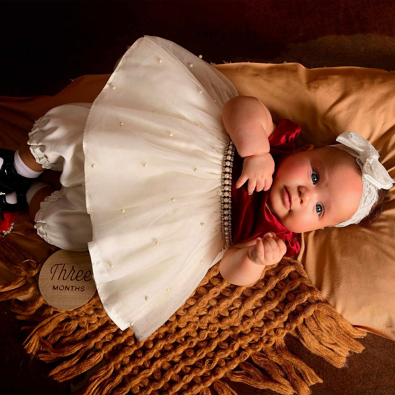 Delanis is registered to the contest to win money with this photo: baby, infant, child, dress, headband, blanket, pillow, brown_background, white_dress, red_top, black_shoes, white_socks, portrait, laying_down, cute, indoors, three_months, decor, soft_texture, cozy