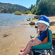 Caydence is registered to the contest to win money with this photo: toddler, child, beach_chair, water, lake, sand, hat, blue_sky, forest, trees, rocks, sunlight, outdoor, nature, summer, swimsuit, footwear, reflection, vacation, relaxation