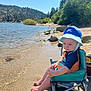 toddler, child, beach_chair, water, lake, sand, hat, blue_sky, forest, trees, rocks, sunlight, outdoor, nature, summer, swimsuit, footwear, reflection, vacation, relaxation